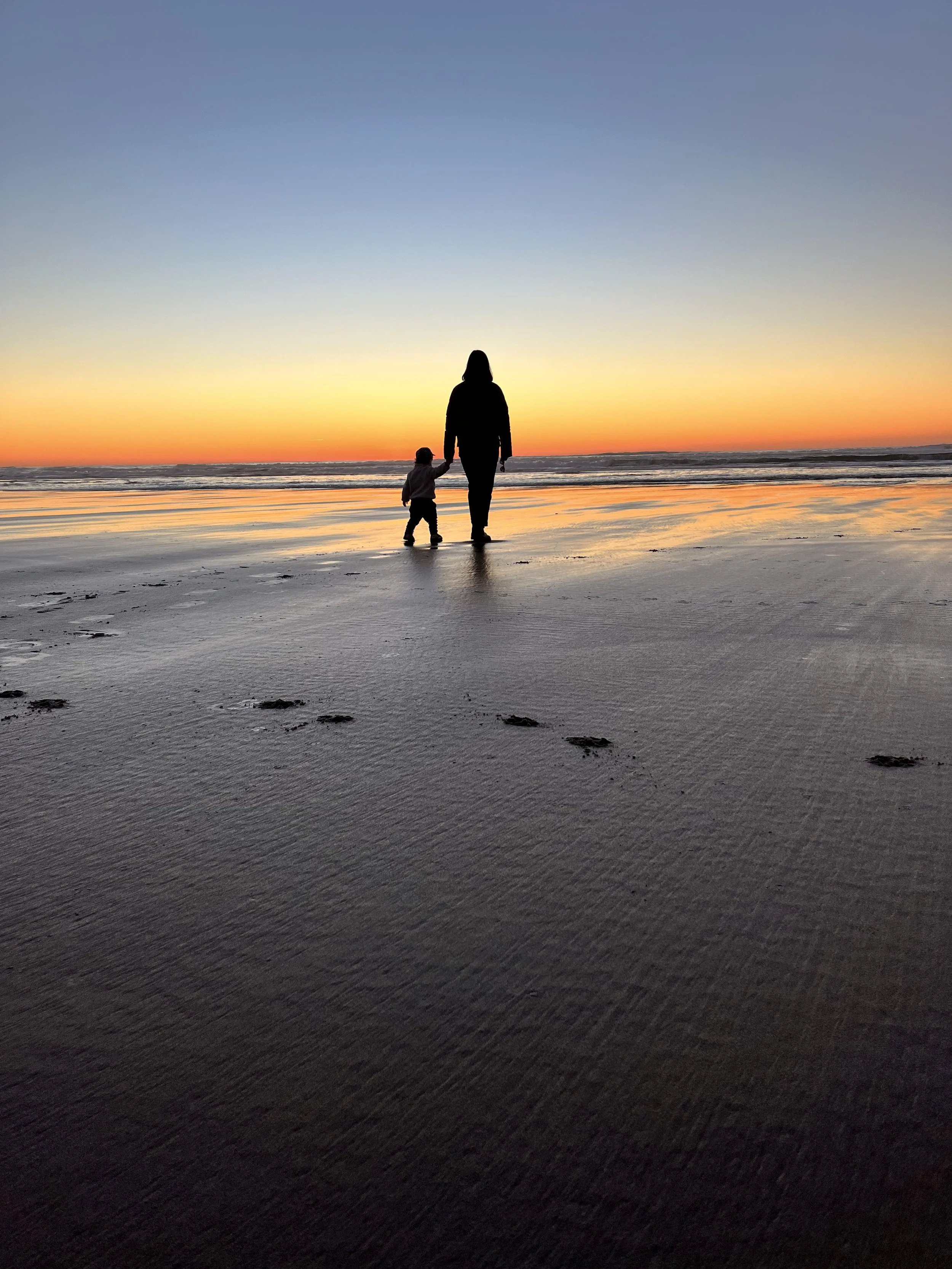 Silhouette of a person and a child holding hands walking on the beach during sunset on the Oregon Coast.