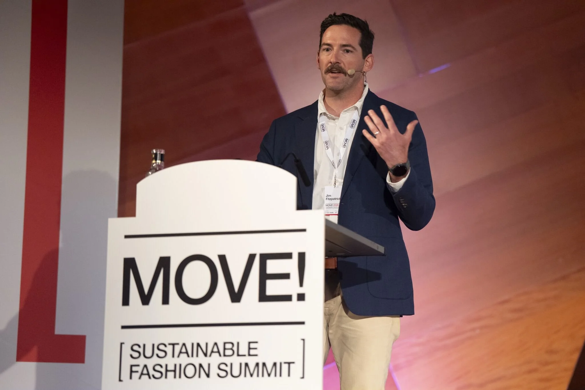 A man with dark hair and a mustache, wearing a navy blazer, white shirt, and beige pants, is speaking at a conference. He has a microphone on his face and a name tag around his neck. In front of him is a white podium with a sign that reads 'MOVE!'. The background features a reddish-brown wall.