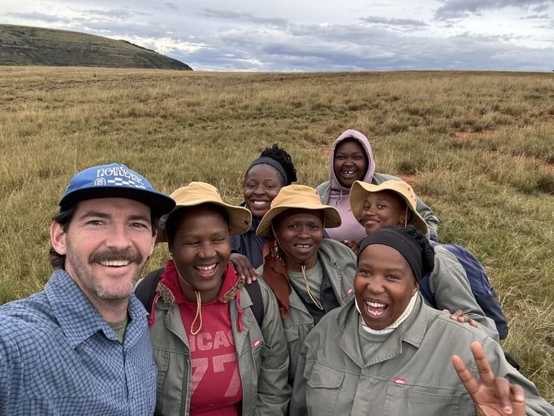 A man taking a selfie with a group of seven smiling women in outdoor field, some wearing hats, under cloudy sky.