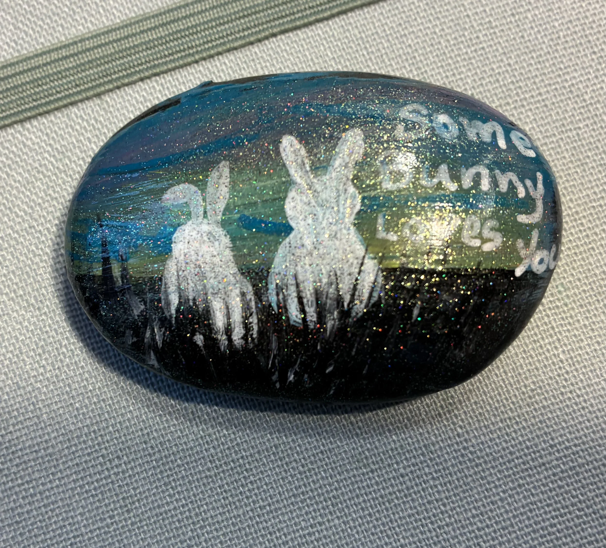 Close-up of a round, decorated glass ornament with glitter, featuring white palm trees and writing, lying on a textured fabric surface.