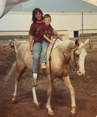 A woman and a young boy riding a light-colored horse together indoors at a riding arena.
