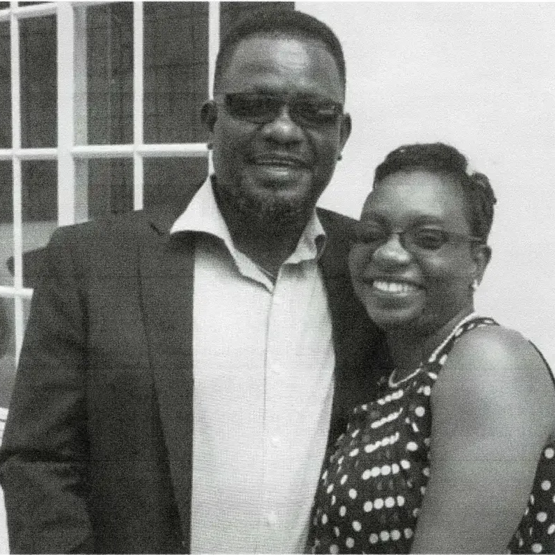 A man and woman smiling and posing together indoors, with the man wearing a suit and glasses, and the woman wearing a polka-dot dress and glasses.