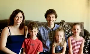 A family of five sitting together on a couch, smiling. The group includes two adults and three children, in a cozy living room.