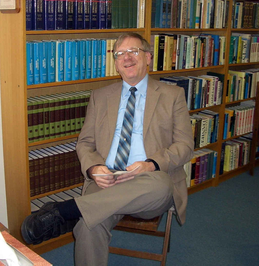 A man in a beige suit and glasses sitting on a chair in front of bookshelves filled with books, smiling and holding a small notebook.