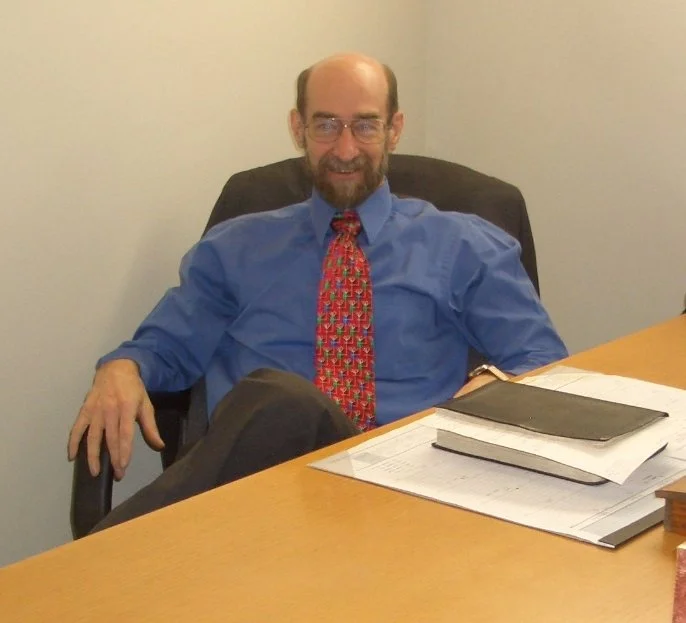 A man with glasses, a beard, and a bald head sitting at a desk, wearing a blue shirt and a festive red tie, with documents and a black folder in front of him.