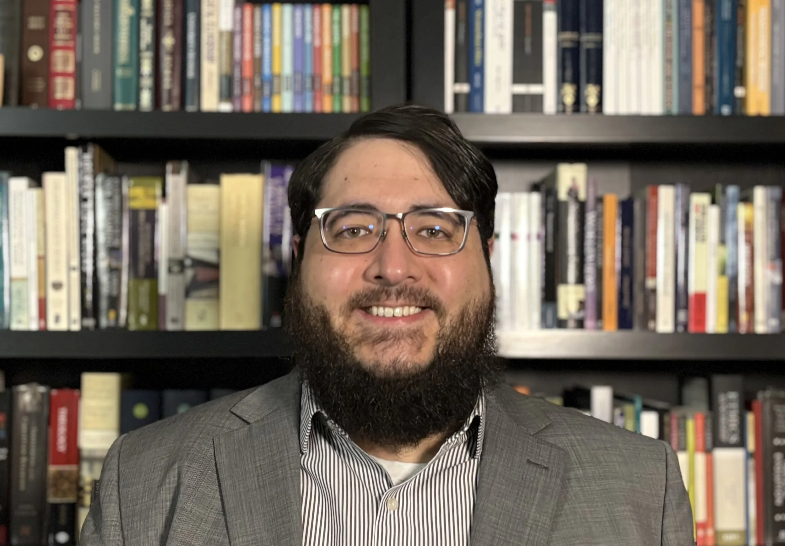 A man with dark hair, glasses, and a beard wearing a gray suit and striped shirt in front of bookshelves filled with colorful books.
