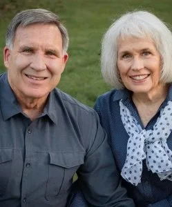 A smiling elderly couple standing outdoors on a grassy field.