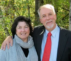 A smiling woman and man standing outdoors among trees, with the woman having dark hair and wearing a light gray scarf, and the man with light hair and a beard, dressed in a dark suit with a red tie.