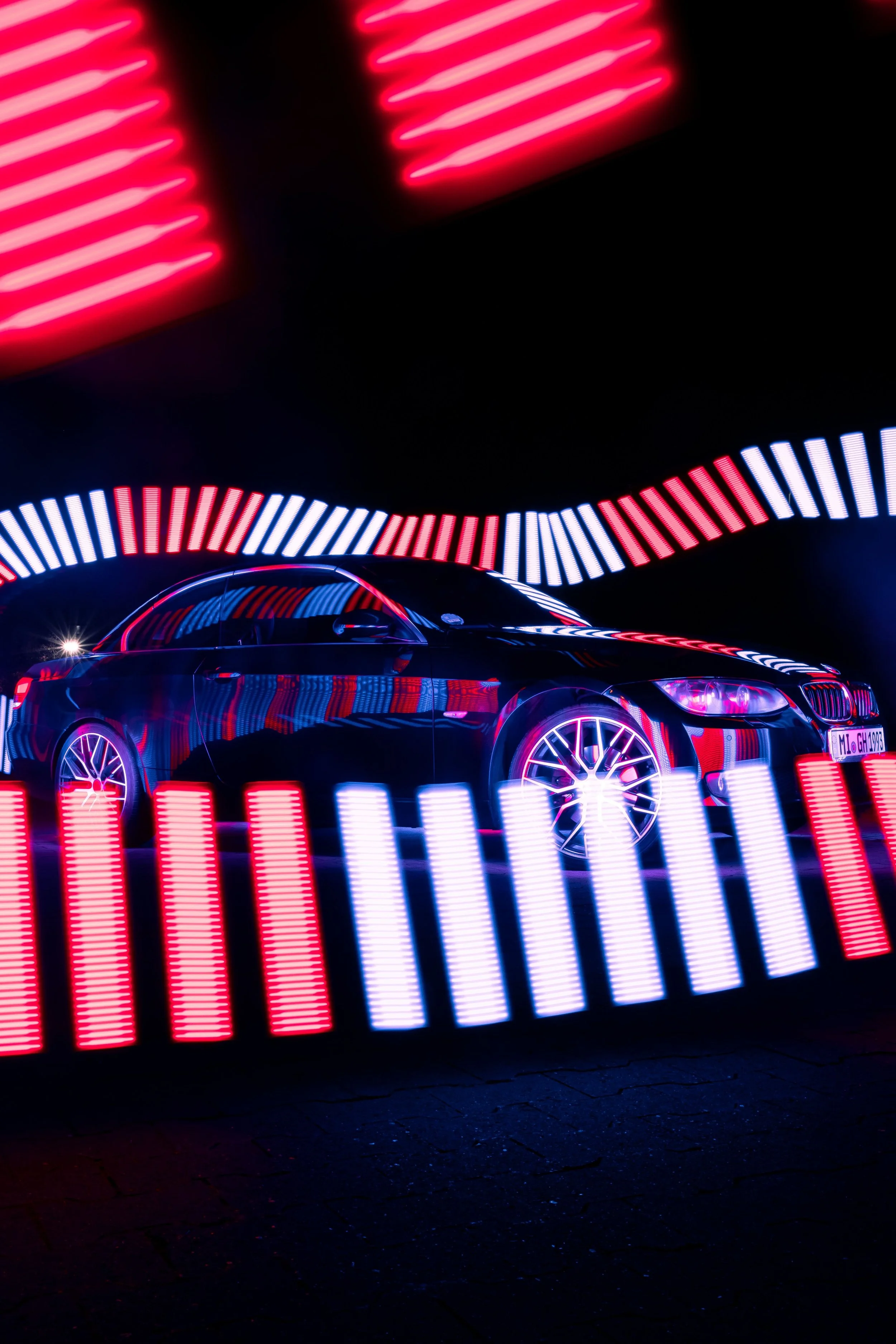Neon light display with red, white, and blue stripes, and a partial view of a black car with reflective surface and illuminated wheels, on a dark street at night.