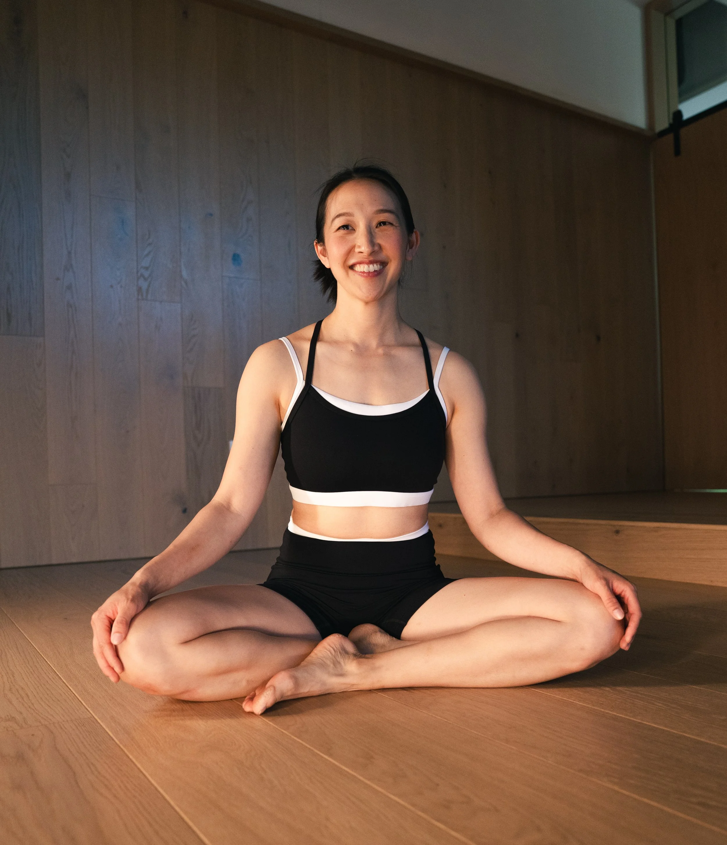 A woman practicing yoga in a seated meditation pose on wooden flooring, smiling, with a beige wall and window in the background.