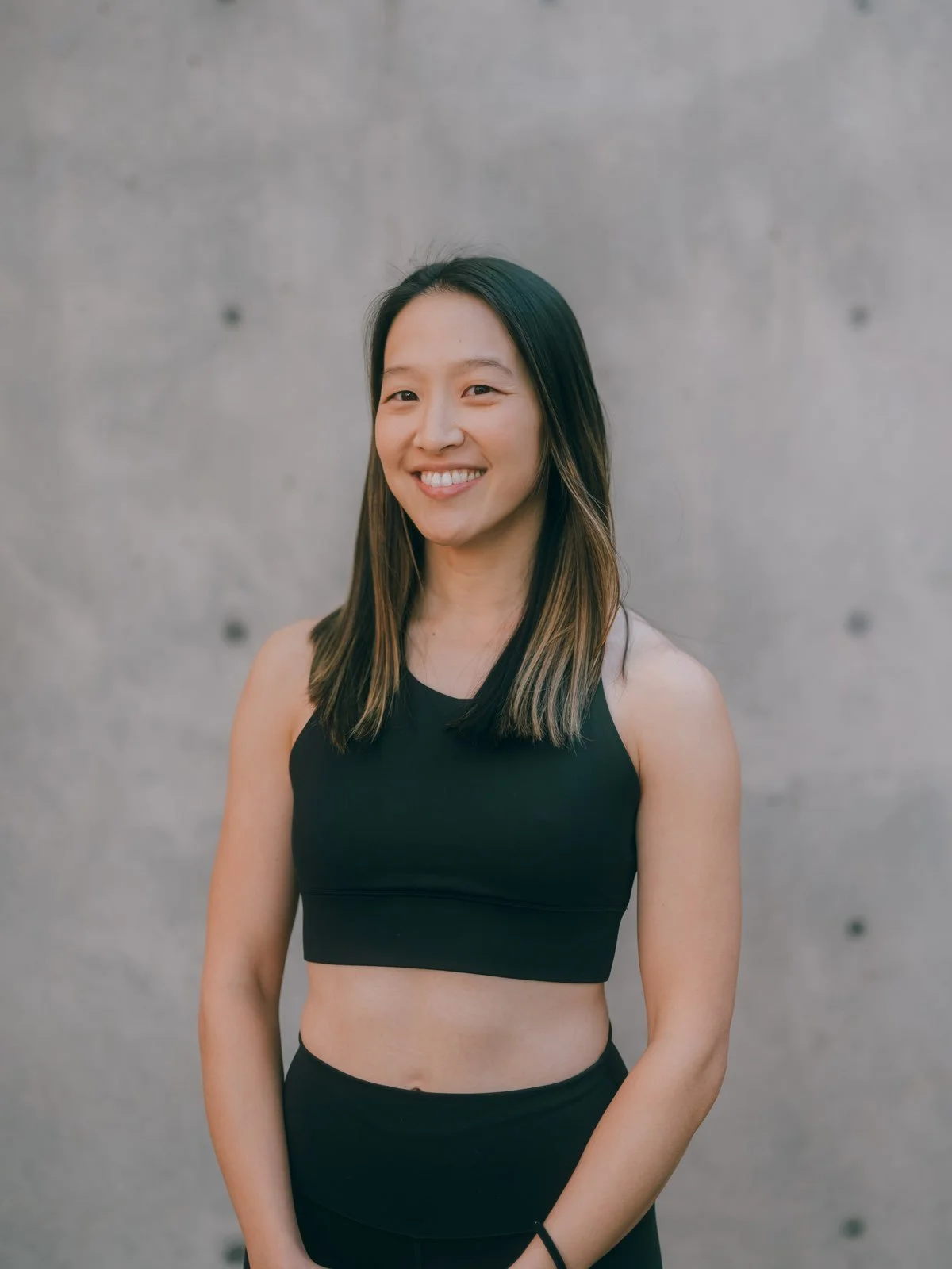 A smiling young Asian woman with shoulder-length dark hair and light brown highlights, wearing a black sleeveless crop top and black workout pants, standing against a gray concrete wall.