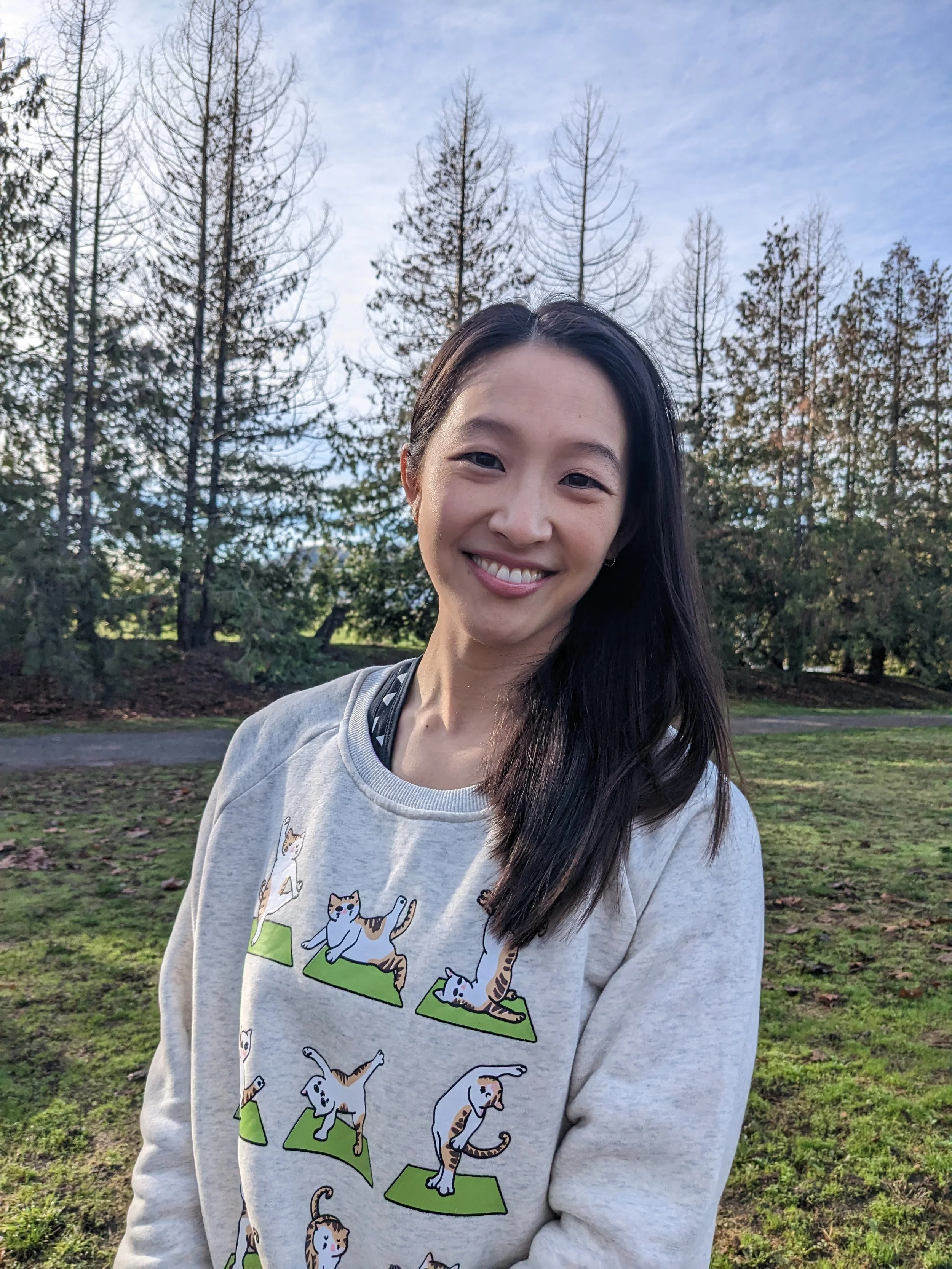 A woman smiling outdoors, wearing a light gray sweatshirt with cartoon cats doing yoga poses, standing on green mats, in a park with trees and blue sky in the background.