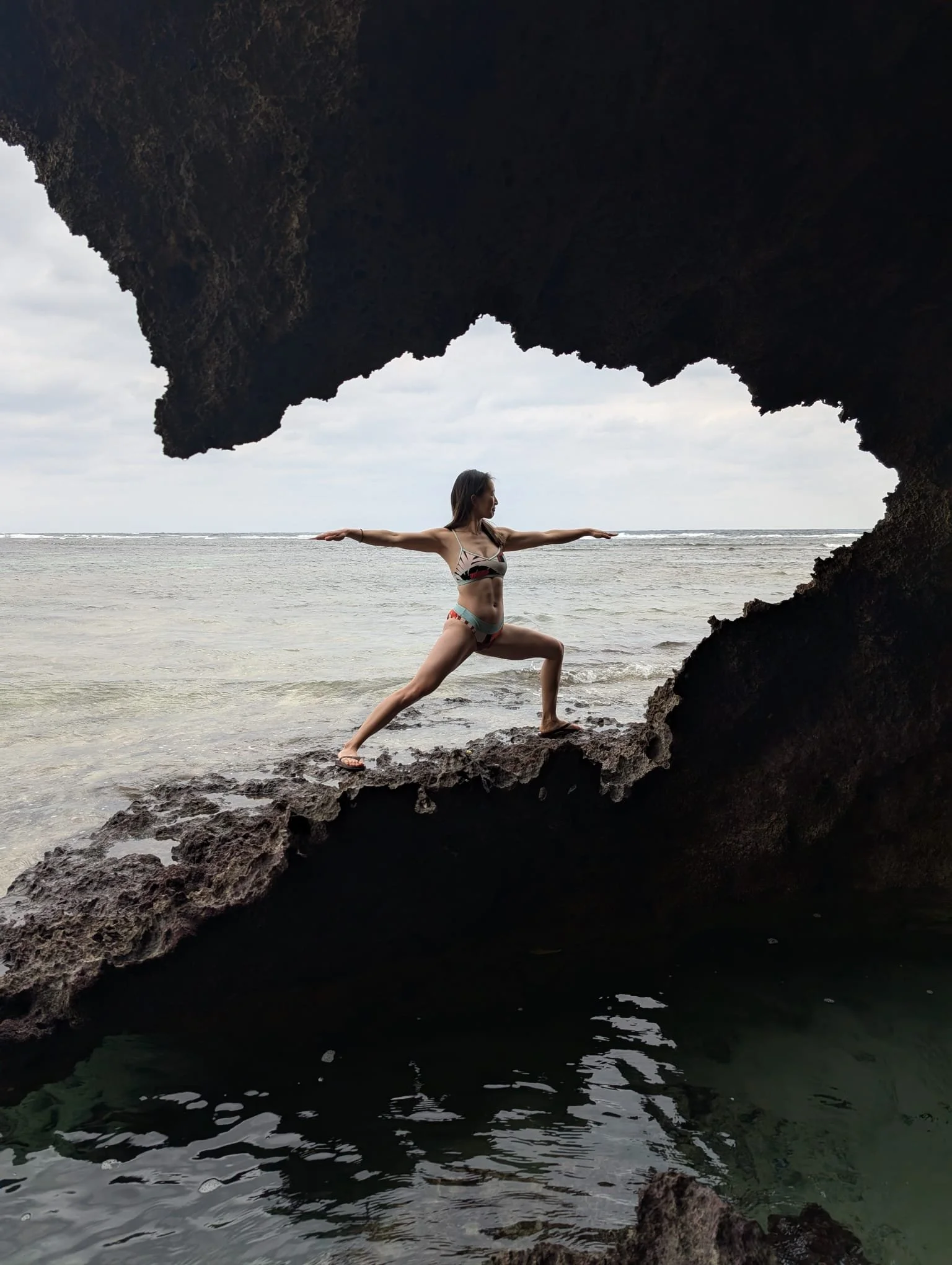 A woman practicing yoga in a lunge pose on rocks at the beach, seen through a natural rock frame with ocean and cloudy sky in the background.
