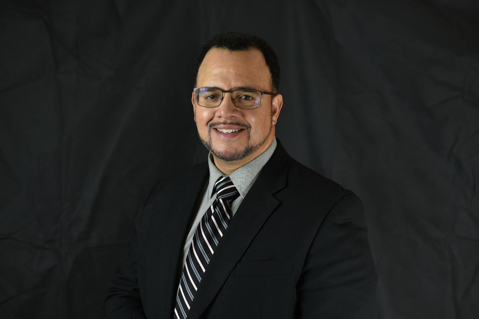 Portrait of a smiling man wearing glasses, a dark suit, a light shirt, and a striped tie, against a black background.