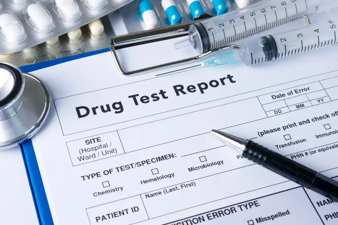 A drug test report form on a clipboard with a pen, syringes, pills, and a stethoscope nearby.
