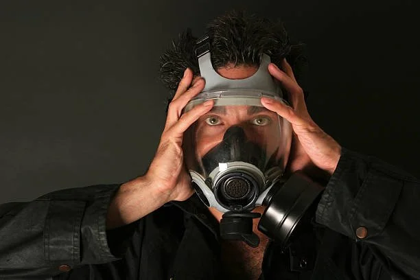 A man wearing a black jacket and a protective mask and goggles, holding his head with both hands, against a dark background.