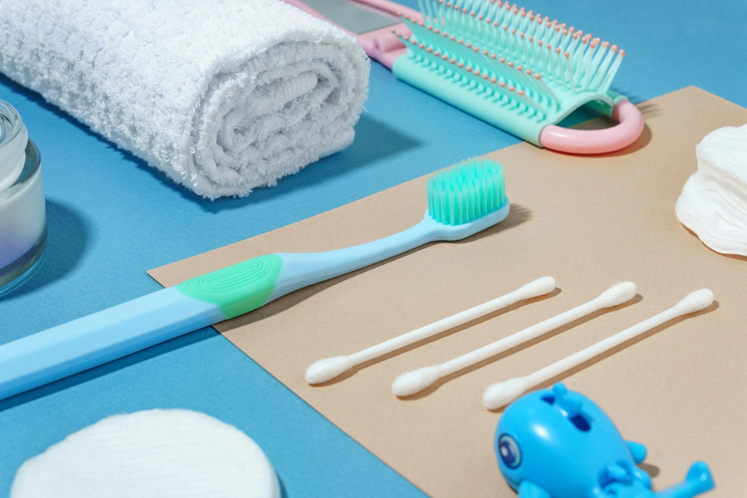 Personal Care & Daily Living - A collection of bath and skincare items including a white towel, a hairbrush, cotton swabs, cotton pads, and cotton balls on a multicoloured background