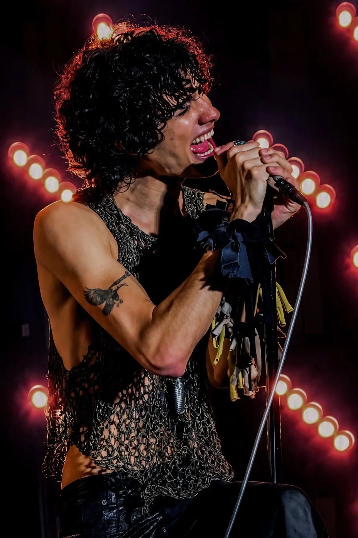 Sombr singing into a microphone in profile with warm red stage lights behind him.