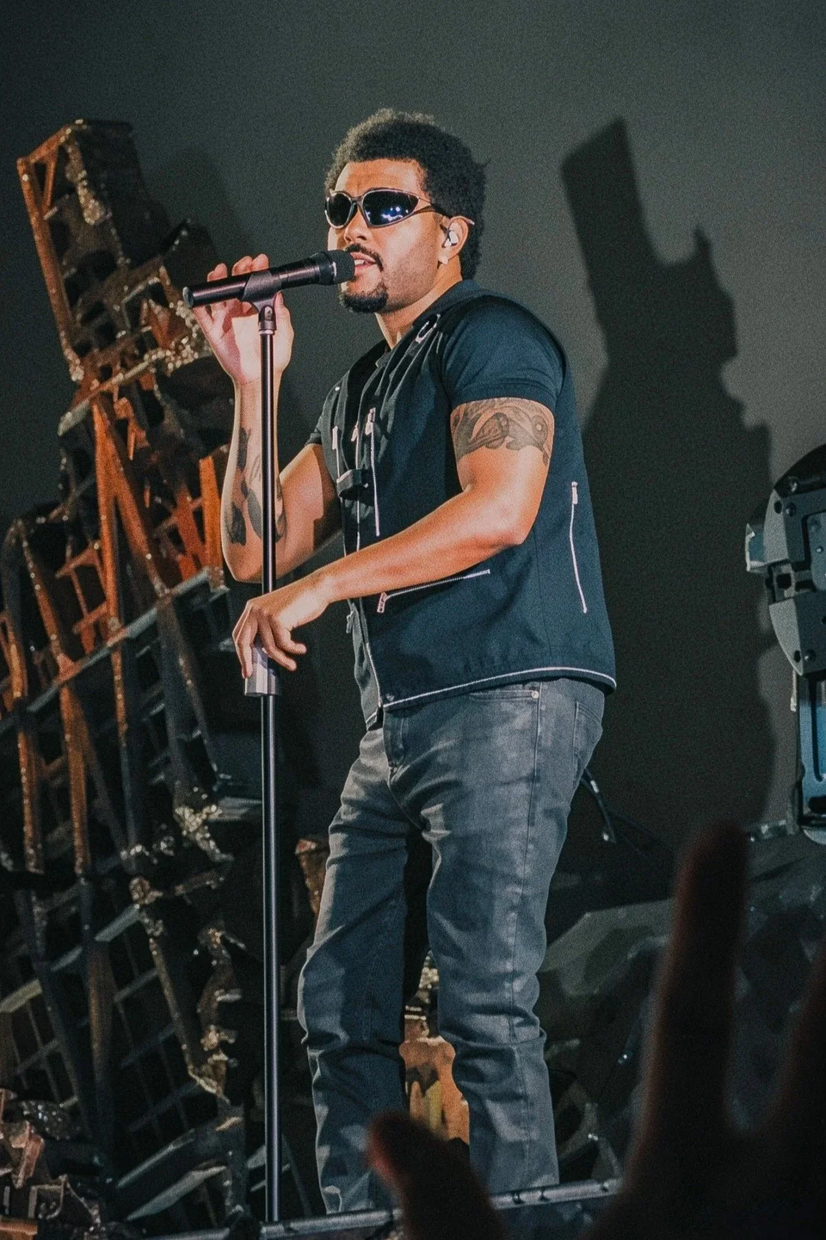 The Weeknd performing in sunglasses beside stage scenery and a dramatic shadow.