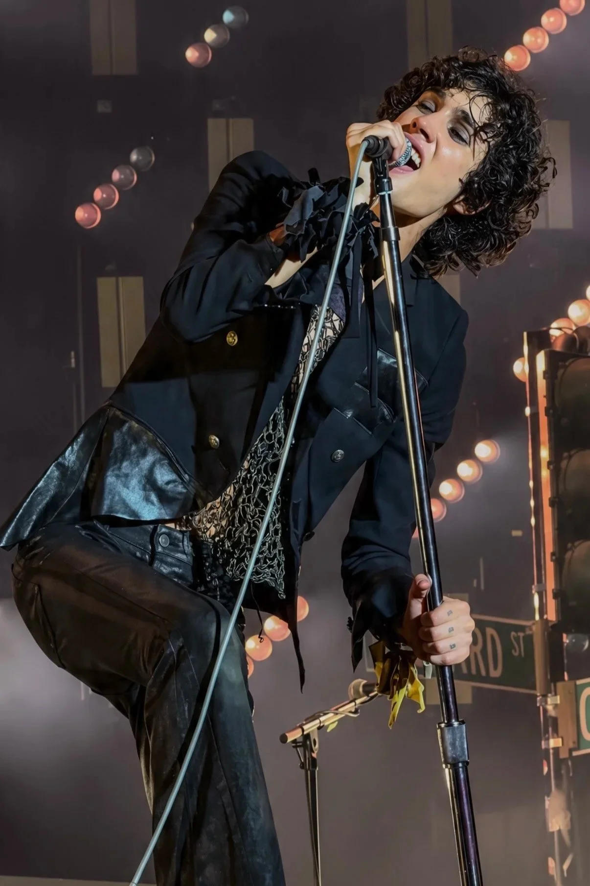 Sombr leaning into a microphone stand while performing in a dark jacket.