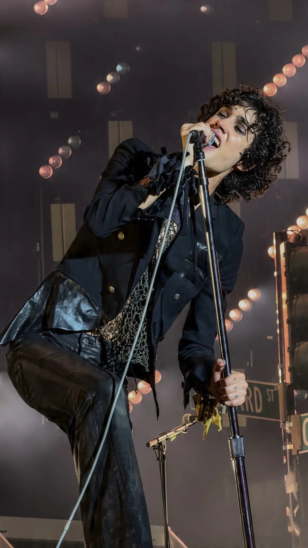 Sombr leaning into a microphone stand while performing in a dark jacket.
