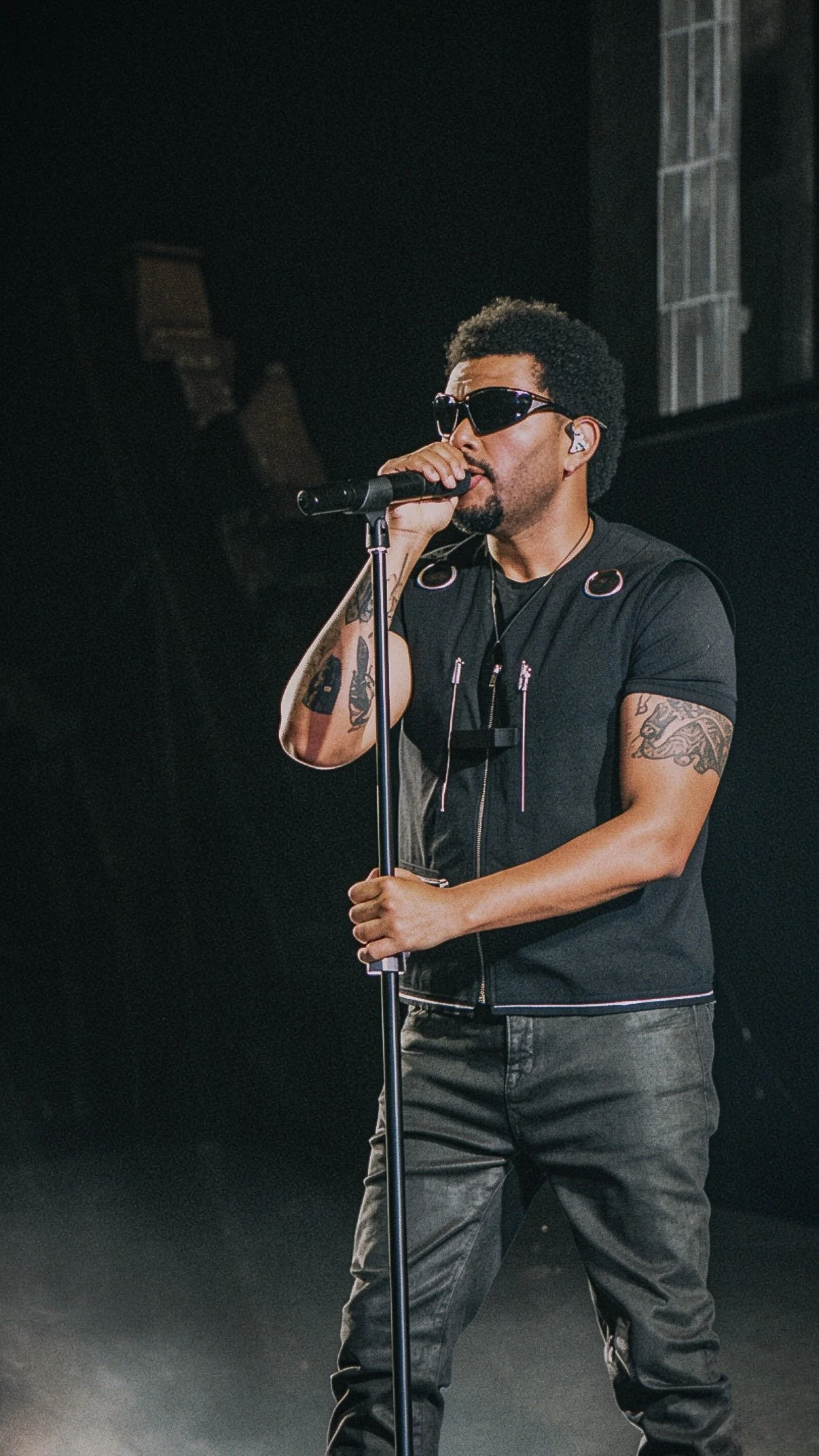 The Weeknd performing at a microphone stand wearing sunglasses on a dark stage.