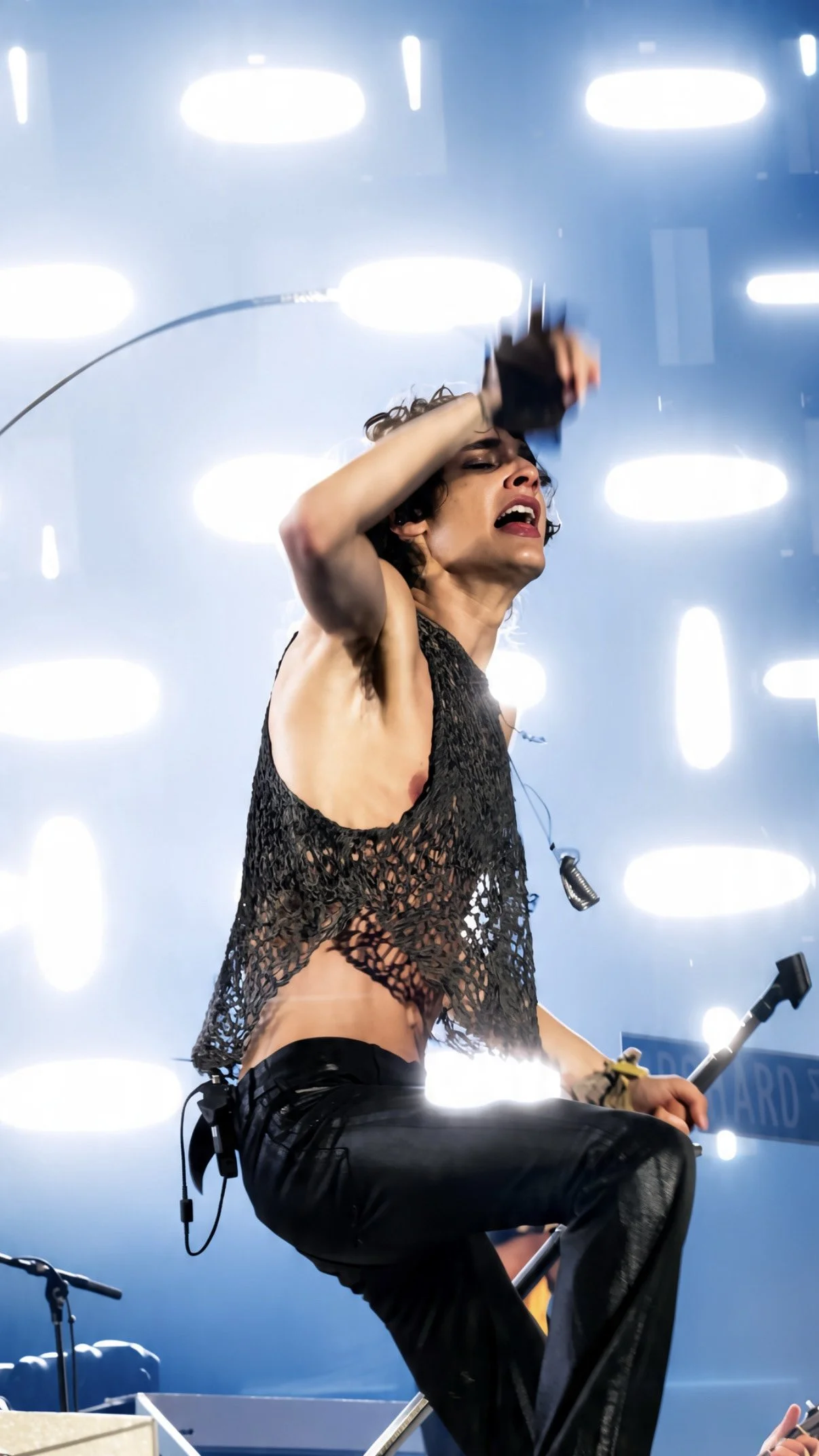 Sombr performing with one knee raised against bright white and blue stage lights.