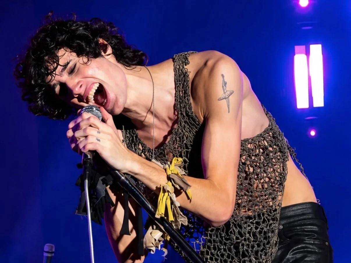 Sombr leaning into the microphone during a live performance under blue stage lights.
