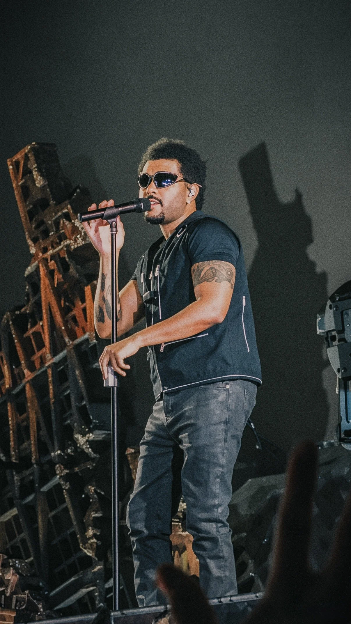 The Weeknd performing in sunglasses beside stage scenery and a dramatic shadow.