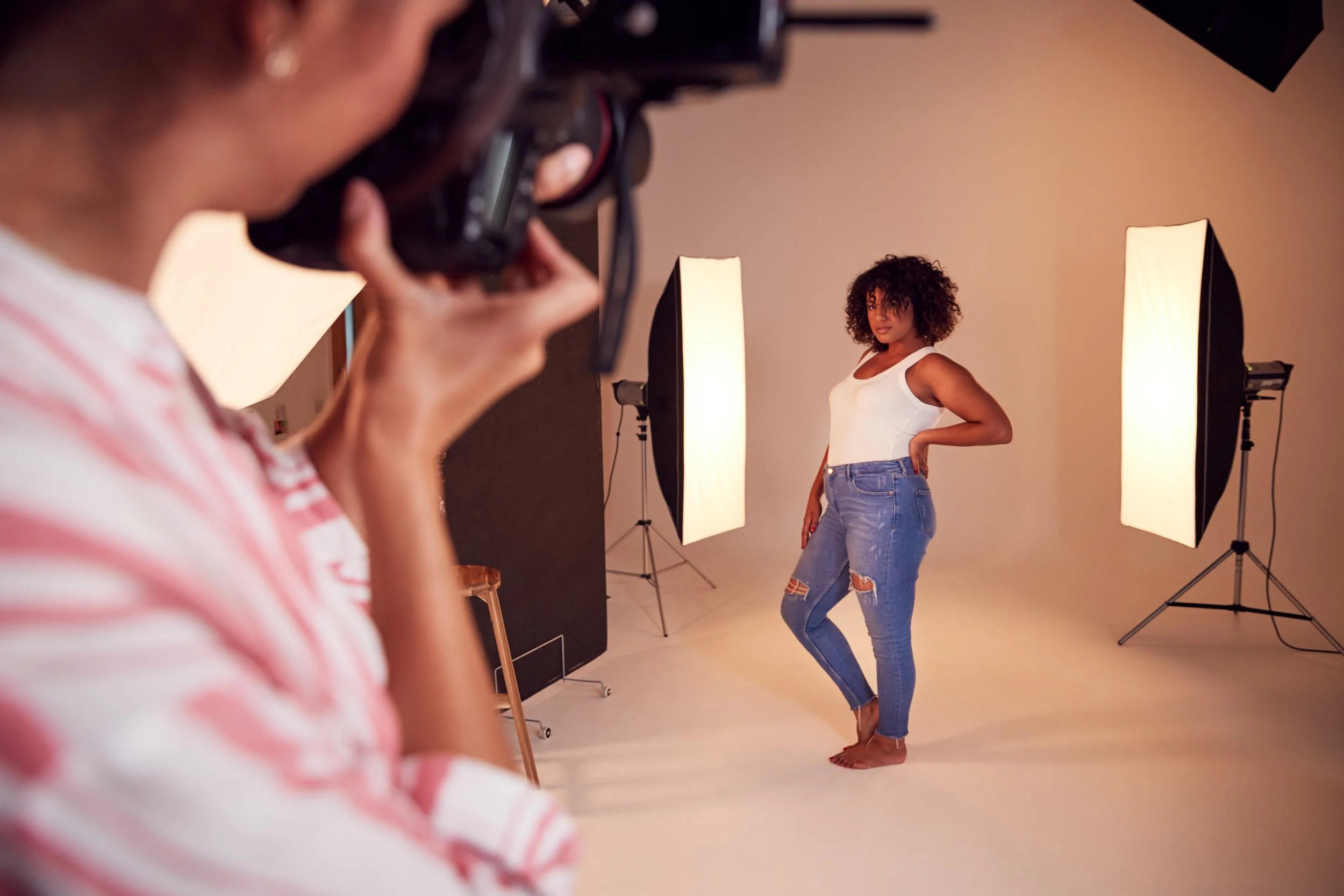 Photographer taking a picture of a woman with curly hair posing in a photo studio with lighting equipment and a beige background.