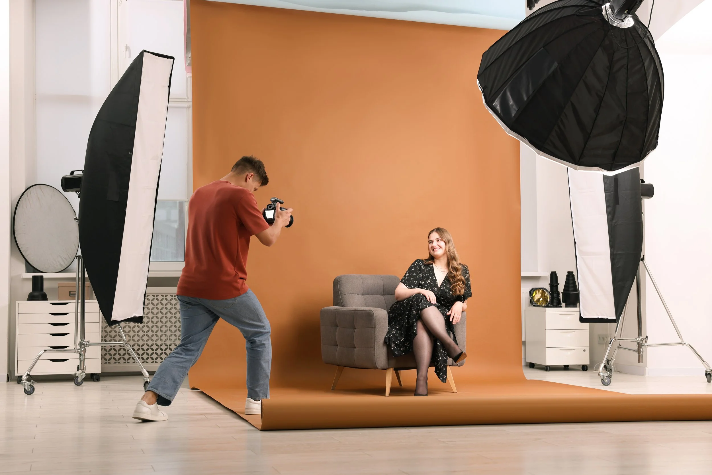 A woman with long hair sitting on a gray armchair in a photography studio, smiling, as a photographer takes her picture. The studio has a brown backdrop, professional lighting equipment, and accessories.