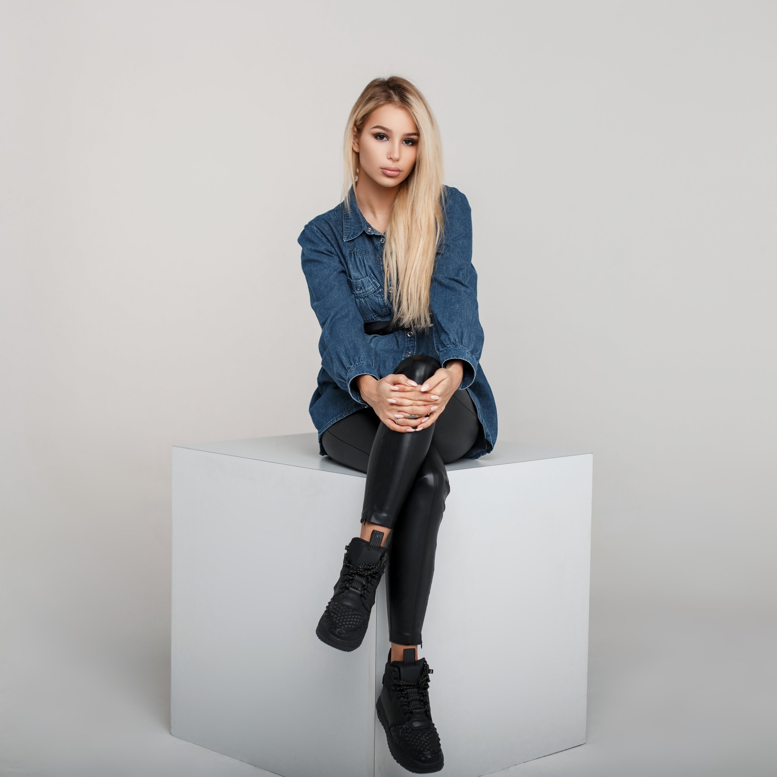 A young woman with long blonde hair sitting on a white platform with her left leg crossed over her right, wearing a denim jacket, black leather pants, and black combat boots, against a plain light gray background.
