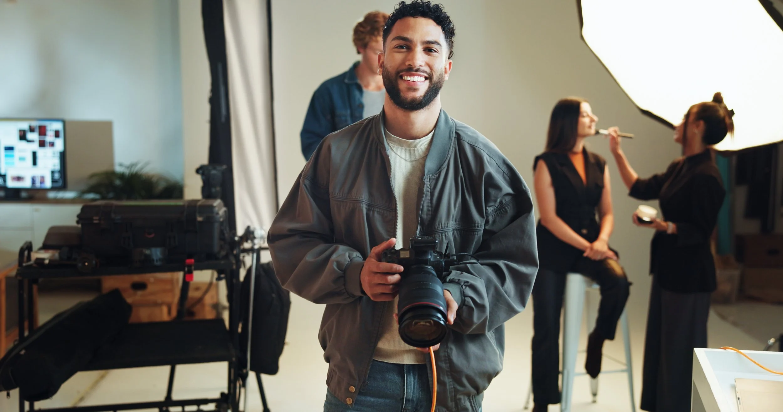A young man with dark curly hair and a beard smiling at the camera, holding a professional camera with a large lens. Behind him, two women are preparing for a photo shoot; one is painting the other's face or makeup, and there is a professional lighting setup and photography equipment in the studio.