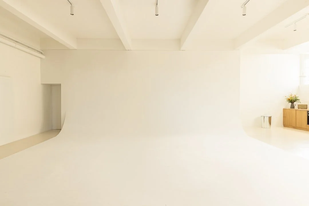 Empty photography studio with a white cyclorama wall and ceiling, minimalist decor, wooden sideboard with houseplants, and a window with sunlight.