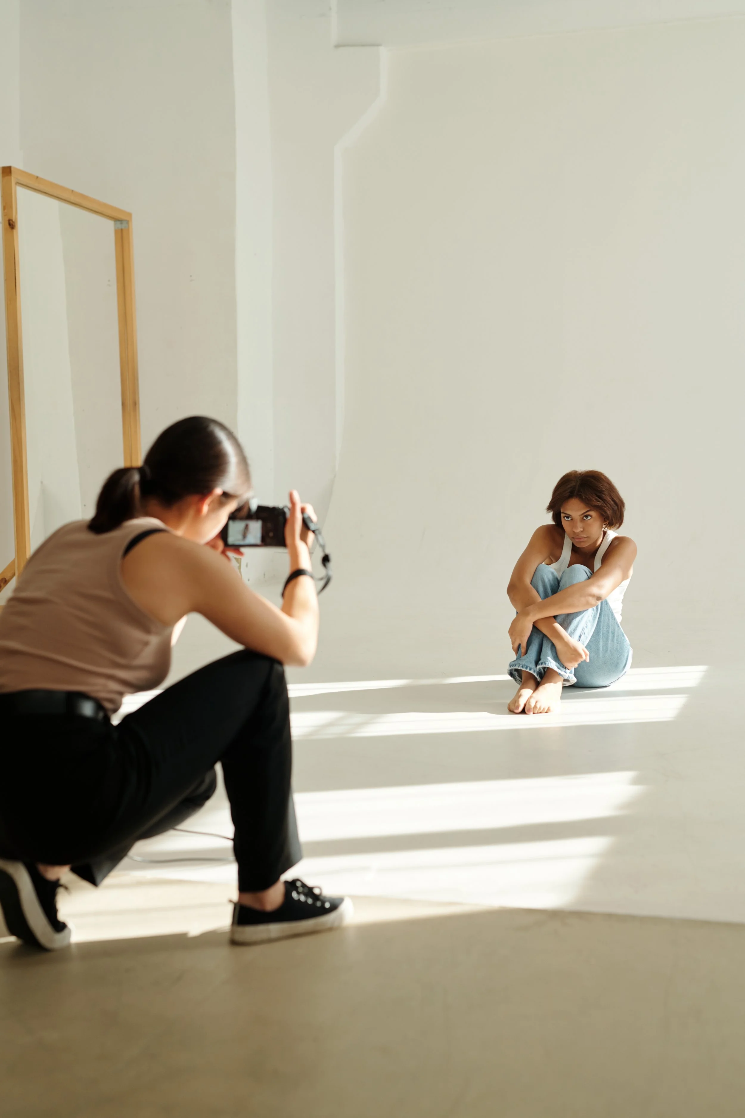 A woman sitting on the floor with her knees drawn up, looking at the camera. Another woman, crouching, is taking her photo. The setting is a minimalist indoor studio with white walls and a wooden frame.
