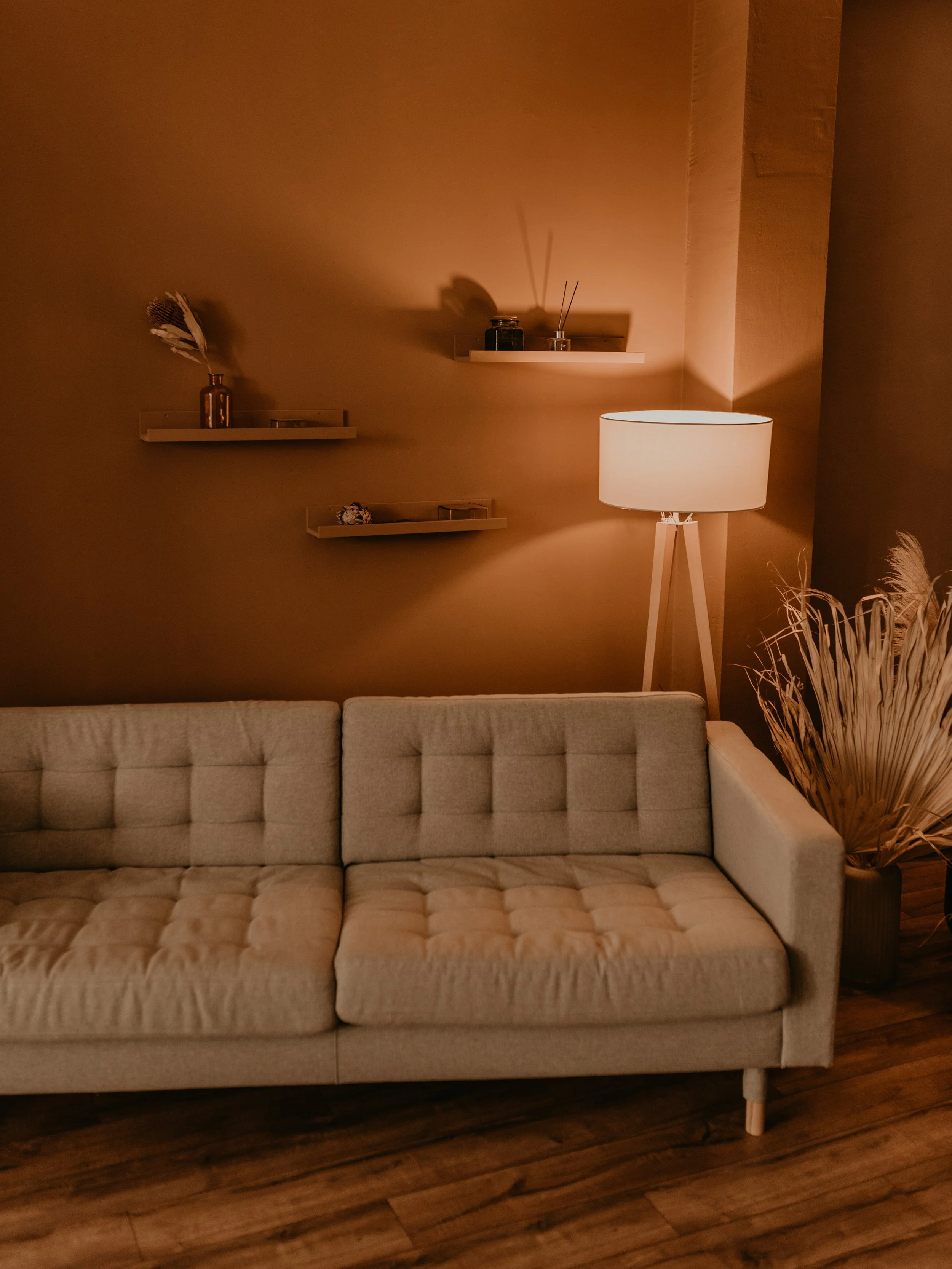 A beige tufted sofa in front of a dark wood floor, with a tall white floor lamp on the right, and wall-mounted shelves with decorative items on the wall behind, in warm ambient lighting.