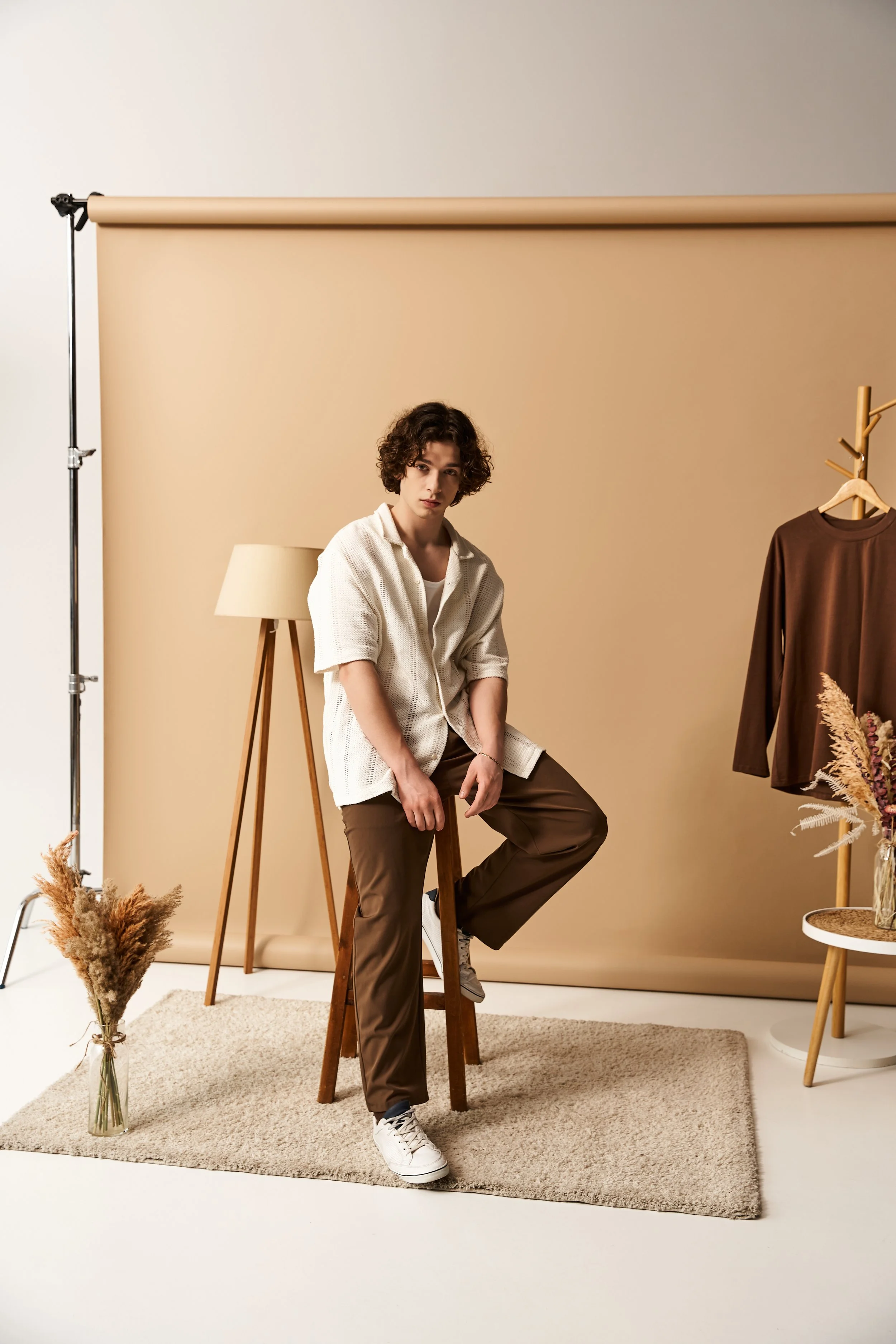 A young man with curly hair sits on a wooden stool in a room with a beige backdrop. He is wearing a white shirt, brown pants, and white sneakers. Behind him is a standing lamp, a clothing rack with a brown shirt, and a small side table with dried plants, all on a beige rug.