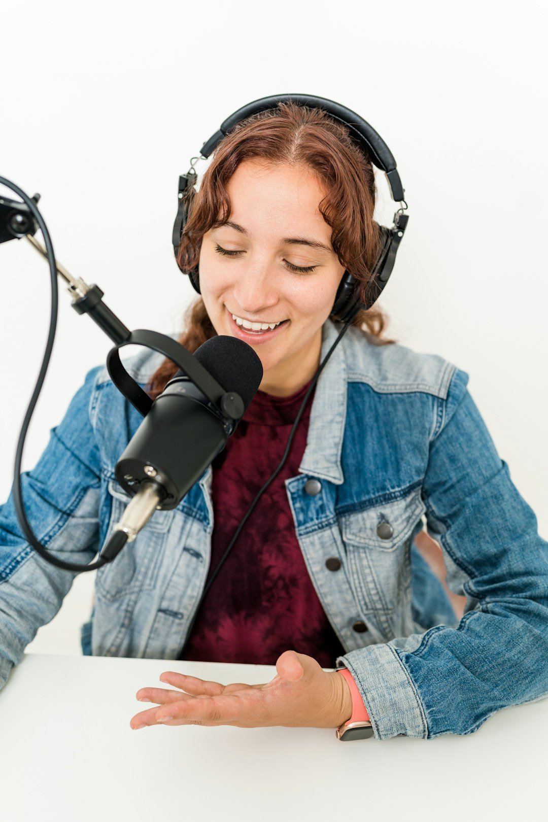 A young woman with curly brown hair wearing headphones and a denim jacket speaking into a microphone in a recording studio.