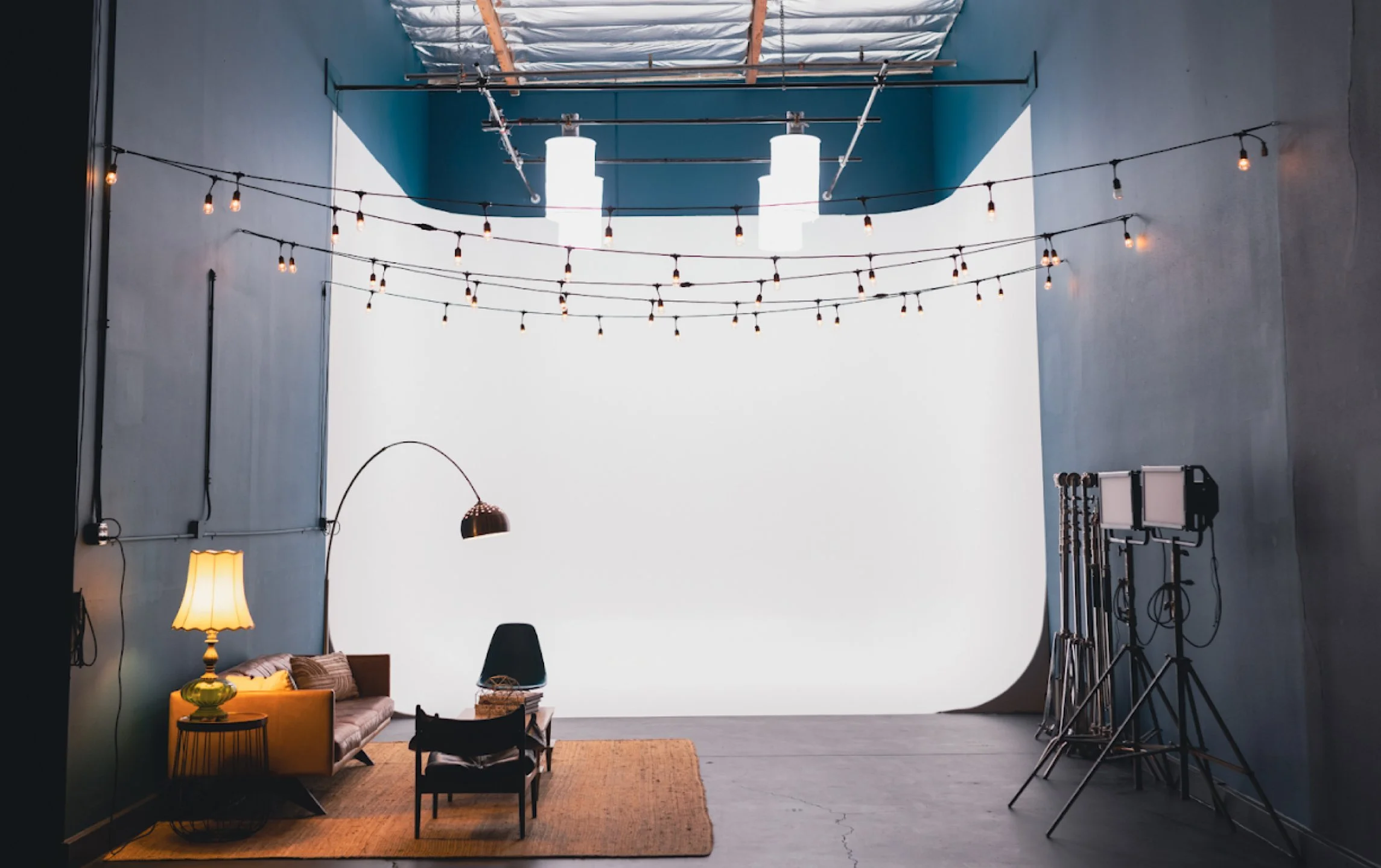 Indoor studio setup with a backdrop, professional lighting equipment, a lounge area with a yellow sofa, a rattan table, a black chair, and various studio accessories.