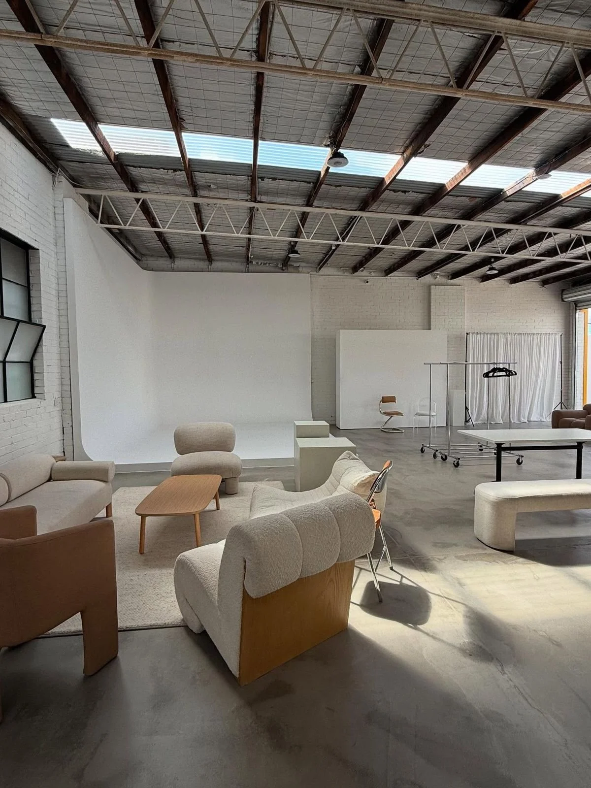 A spacious, industrial-style photo studio or lounge with white brick walls, a high ceiling with exposed beams, and large windows. The foreground features modern beige and white furniture, including armchairs, a sofa, and a wooden coffee table. In the background, there is a white backdrop, a small table, chairs, and a clothing rack with hangers. The overall atmosphere is minimalist and clean.