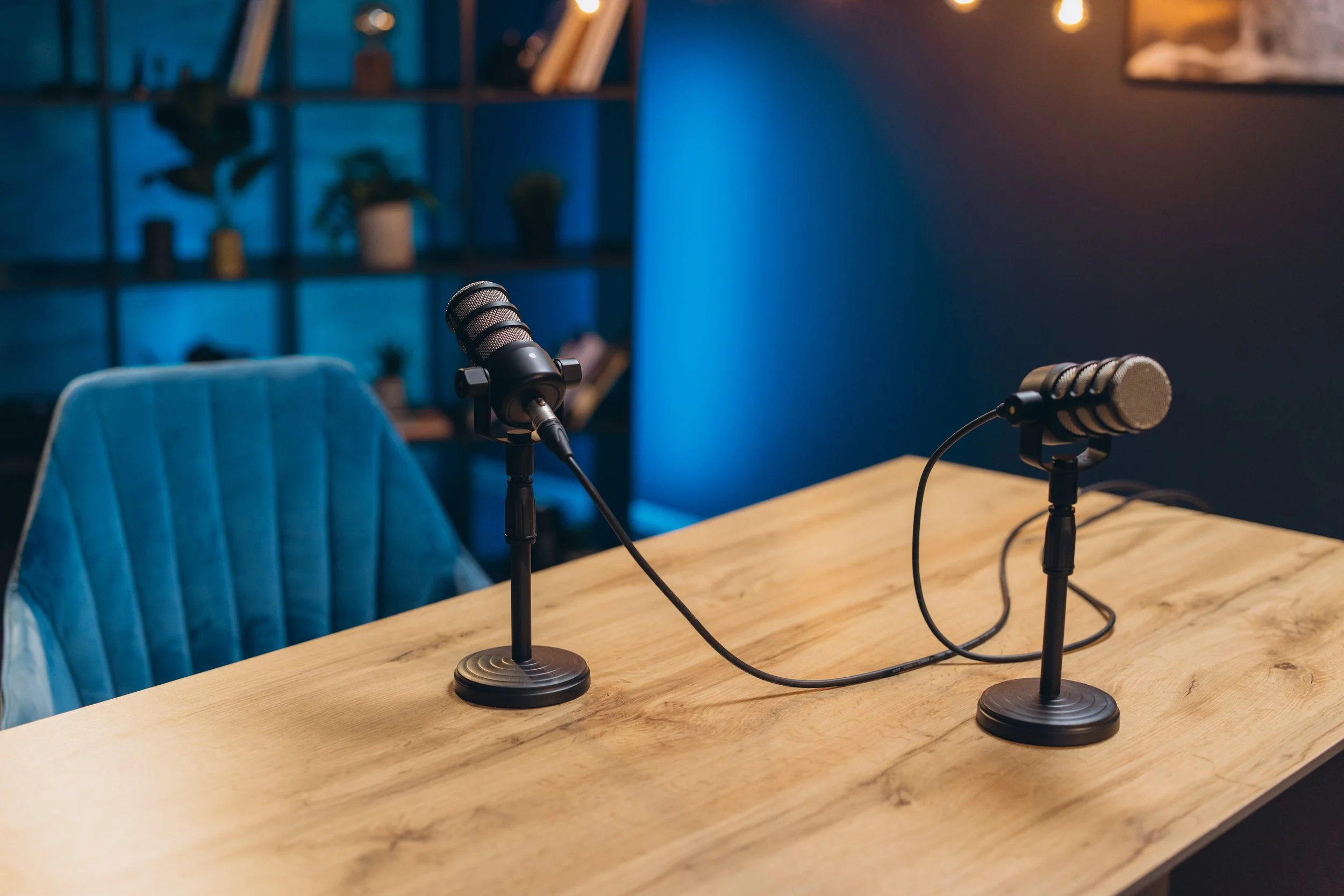 Two microphones on stands positioned on a wooden table, with a blue armchair nearby. In the background, there is a dark-colored wall, a bookshelf with potted plants and books, and warm string lights. The setting appears to be a podcast or recording studio.