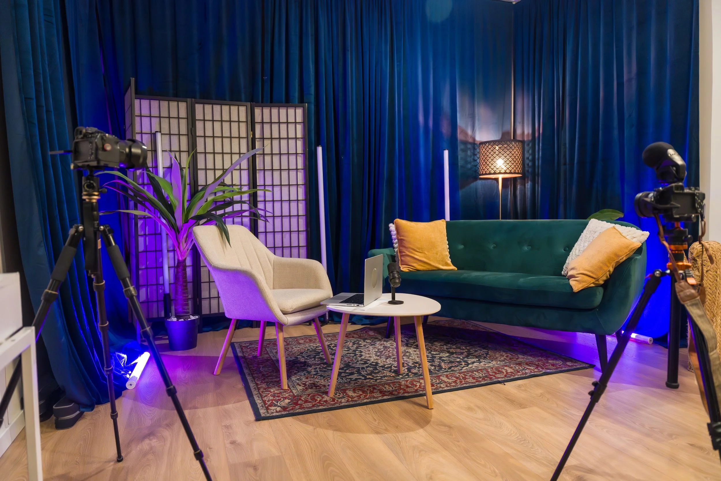 A studio setup with blue curtains, a green sofa with pillows, a white chair, a small table with a laptop and microphone, two cameras on tripods, and various lighting and decorative elements.