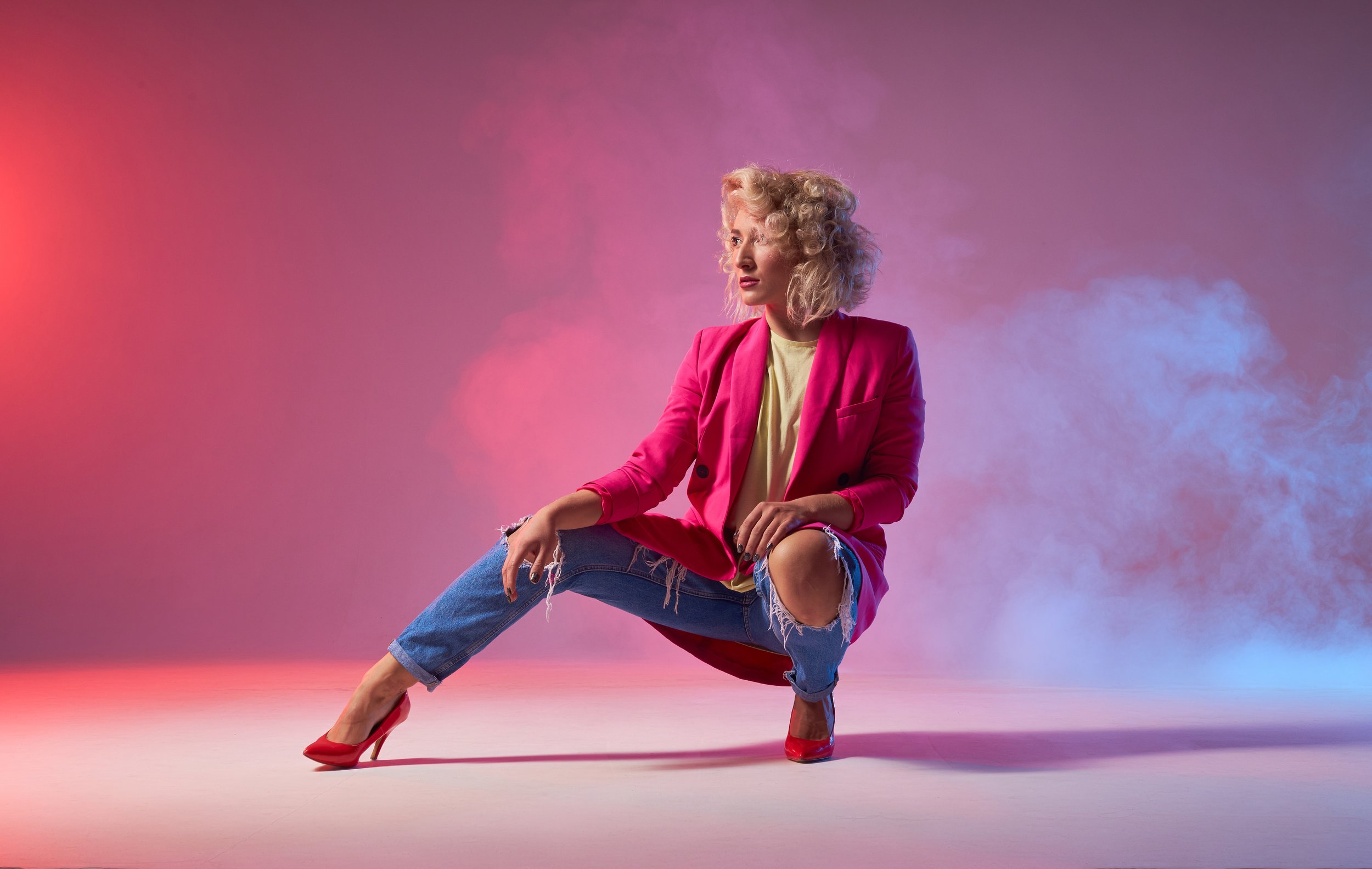 A woman with curly blonde hair wearing a pink blazer, yellow top, ripped blue jeans, and red high heels is posing against a pink and purple gradient background with colored smoke.