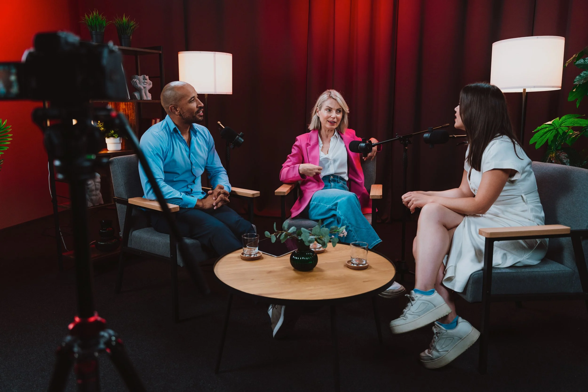 Three people sitting in a semi-circle engaged in conversation in a room with dark red walls. There are two women and a man. The woman on the right is wearing a white dress and sneakers, the woman in the middle is wearing a pink blazer and jeans, and the man is wearing a blue shirt. There is a camera on a tripod in the foreground, and the setting appears to be a podcast or interview studio with microphones, lamps, and plants.