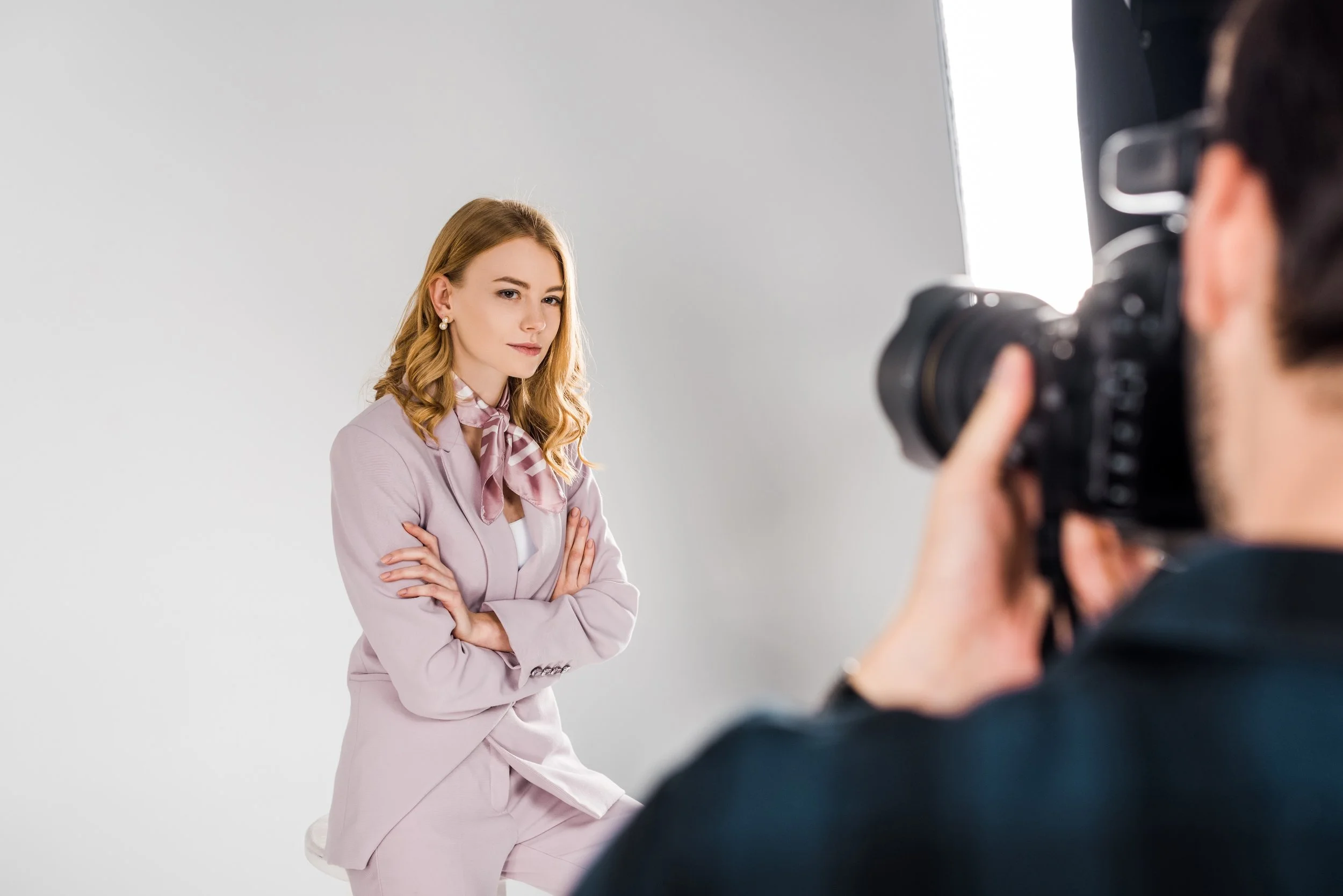 A woman with long red hair wearing a light pink suit and decorative earrings poses with crossed arms in front of a gray background, photographed by a person using a professional camera.