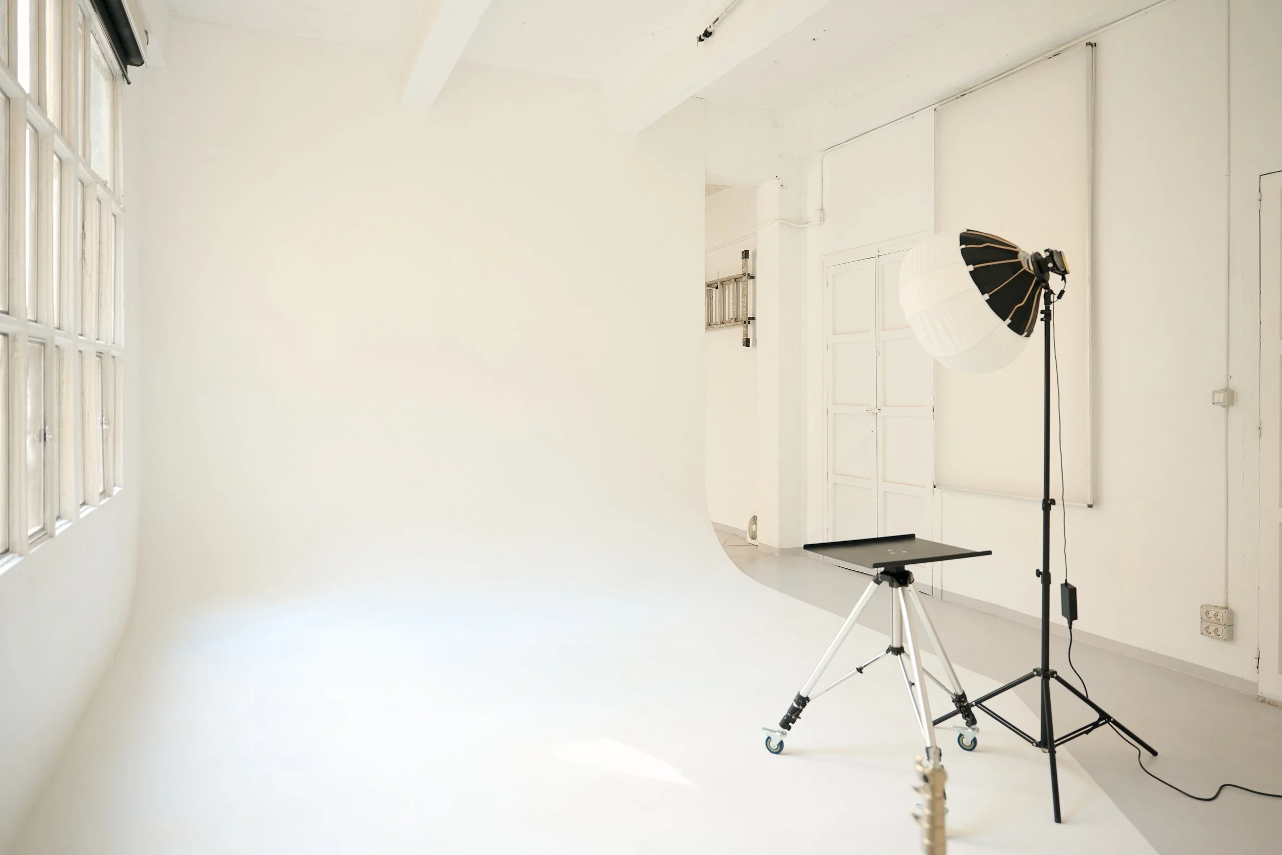 An empty photography studio with a white wall, large window with glass blocks, a softbox light on a stand, and a camera on a tripod.