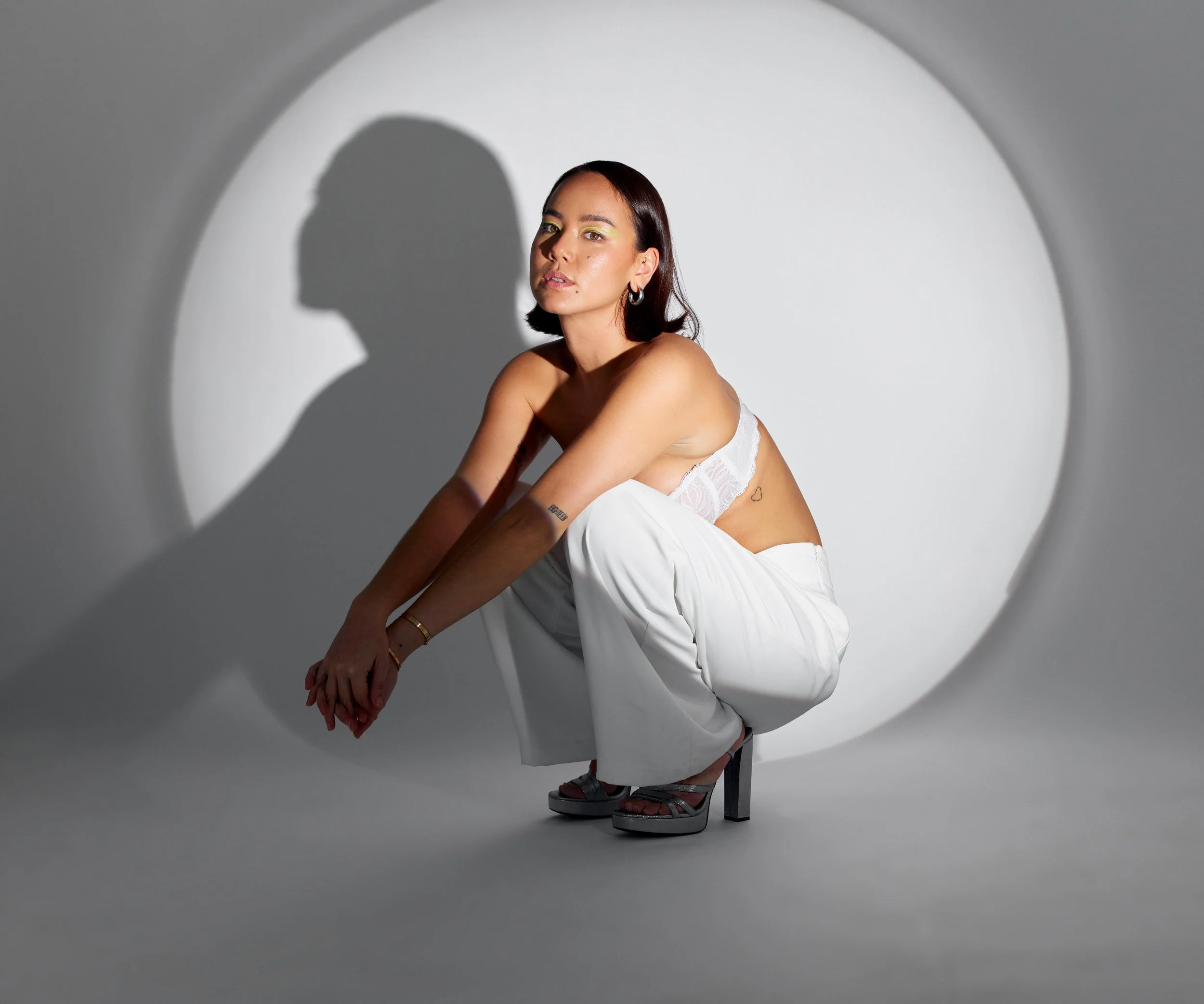 A woman with short dark hair wearing a white dress and silver platform sandals, sitting on a chair with her arms crossed, casting a shadow on a white wall in a minimalistic setting.