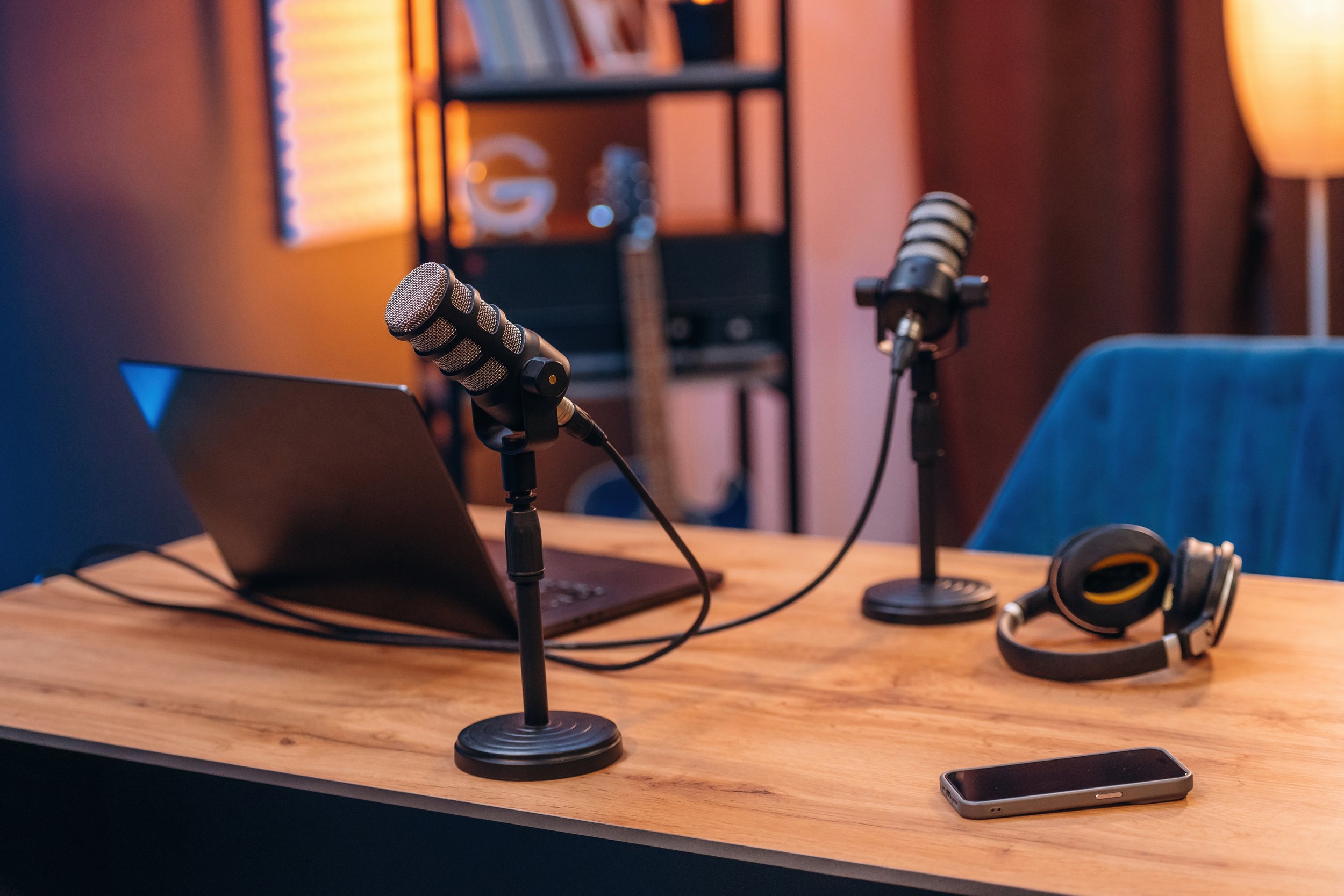 A wooden table with a laptop, two microphones on stands, a pair of headphones, and a smartphone on it, in a room with soft orange lighting.