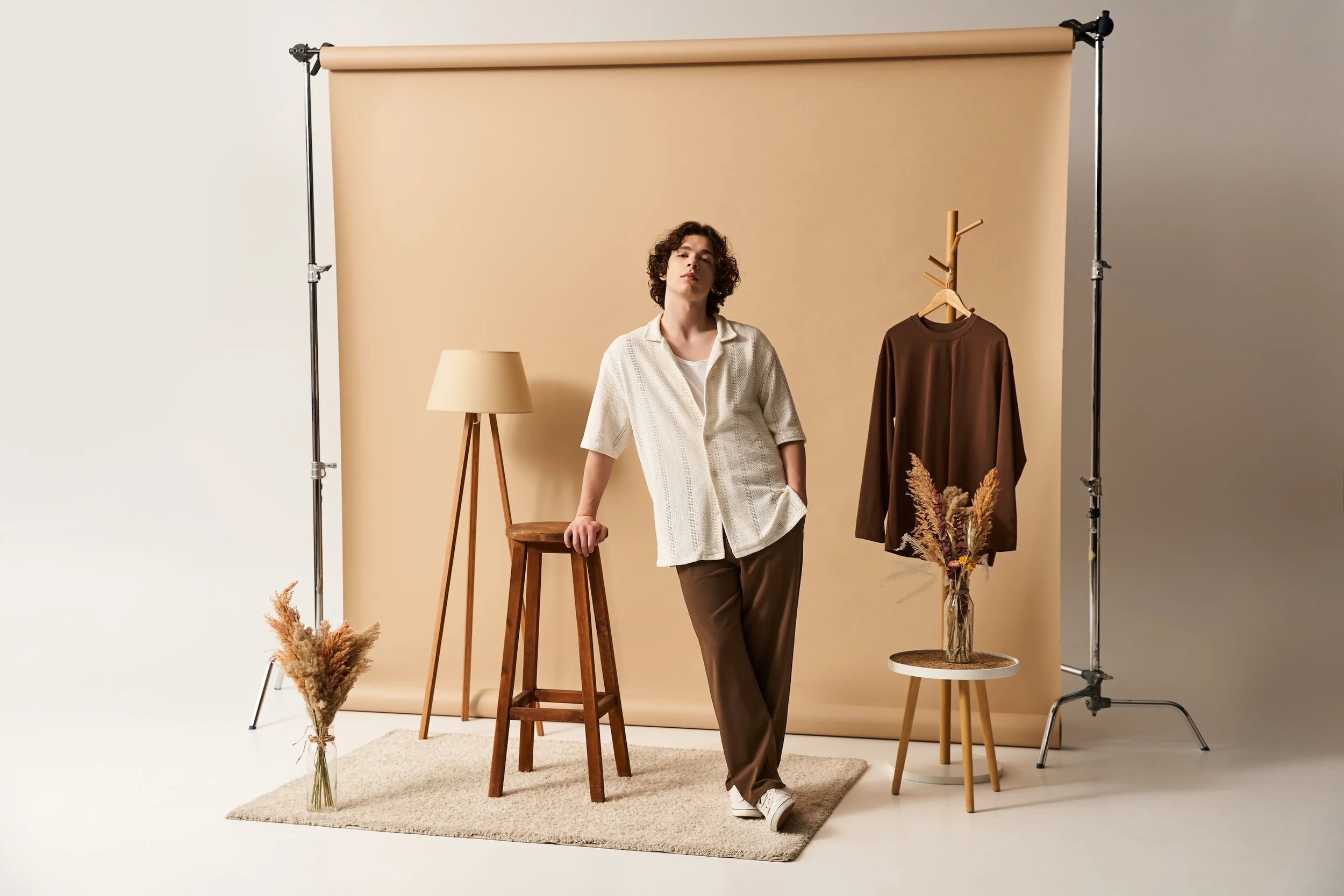 A young man with curly hair standing in a photo studio with a beige backdrop, a wooden stool, a beige floor lamp, a brown shirt on a hanger, and dried flowers in a glass vase.