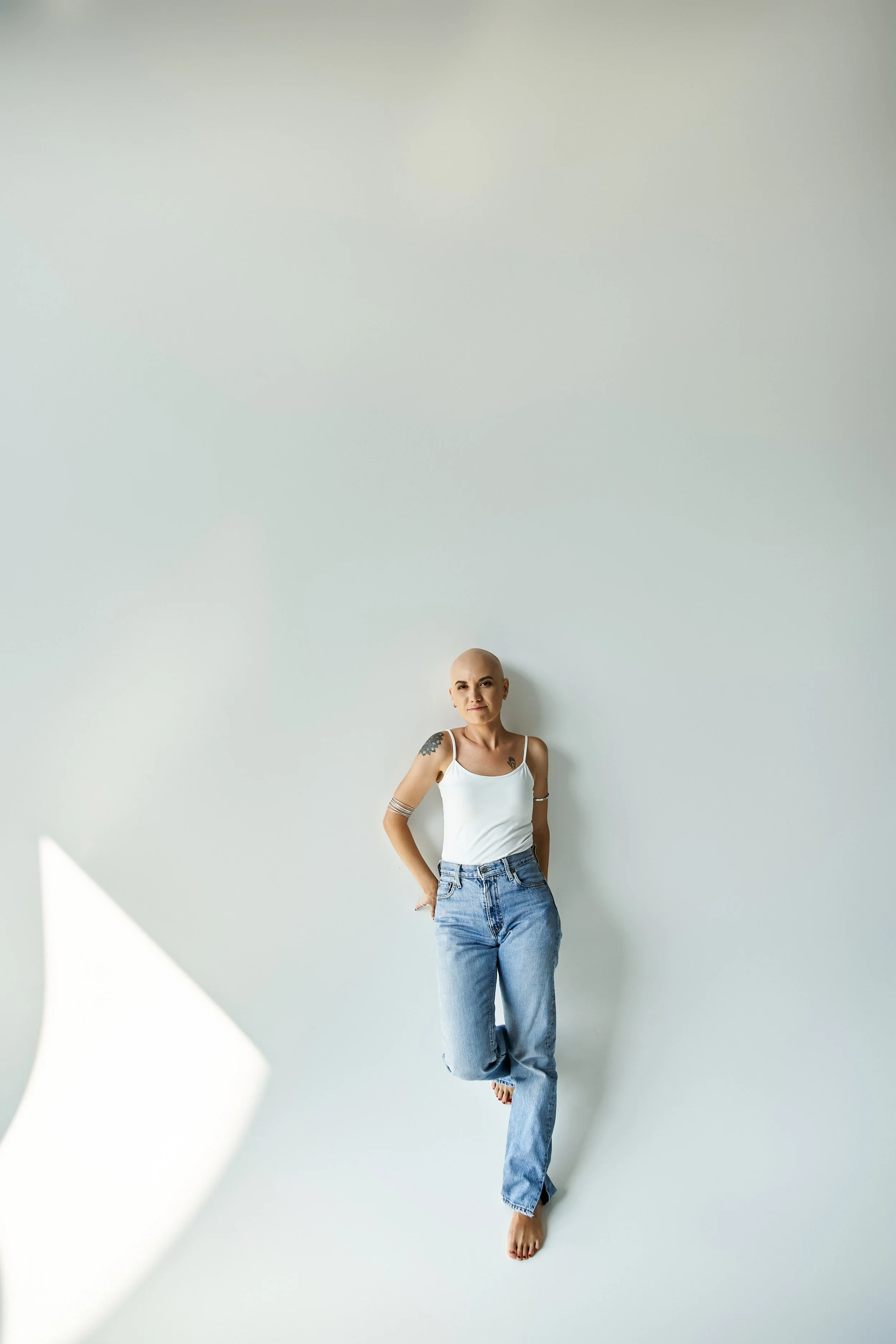 A woman with a shaved head and tattoos on her arms standing against a plain white wall, wearing a white tank top and blue jeans.